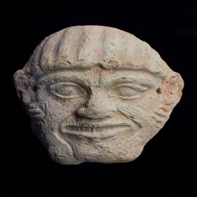 Mask. Hawawa face, vertically ribbed hair or hat, large ears, facial decoration on cheeks and jaws. Wide grinning mouth exposes teeth. Two holes pierced in front of ears. Traces of red paint under chin. Clay.; YPM BC 016861