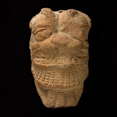 Terracotta figurine of the head of the demon Pazuzu. Pazuzu head and neck. Hair parted in vertical rows with two horns, prominent whiskers and patterned beard extending onto neck in three tresses. Mouth open in growl, exposing rows of sharp teeth and tongue. Hole pierced on top for suspension.; YPM BC 016825